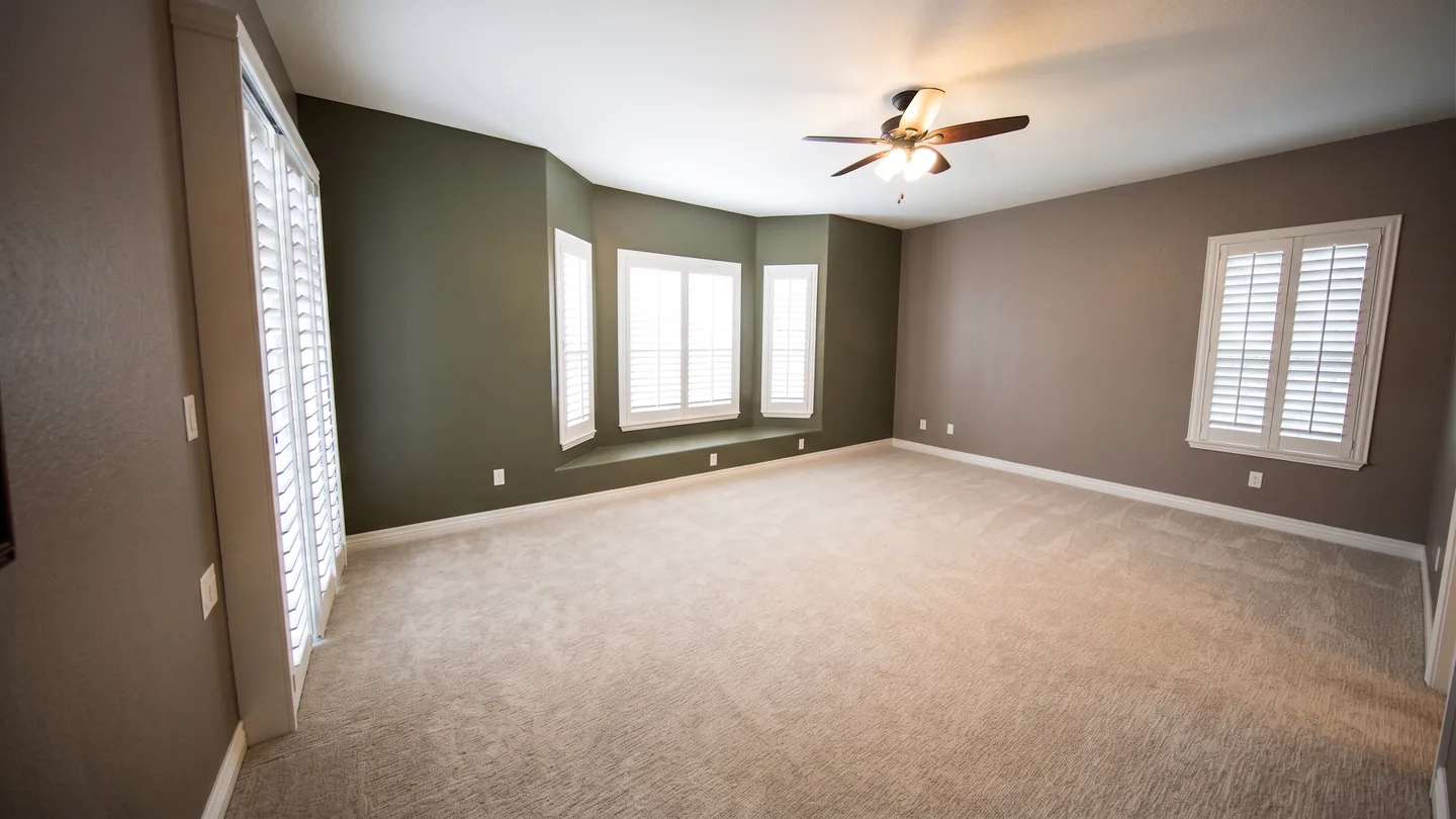 The same bedroom emptied — bay window, plantation shutters, ceiling fan and carpet all where they were