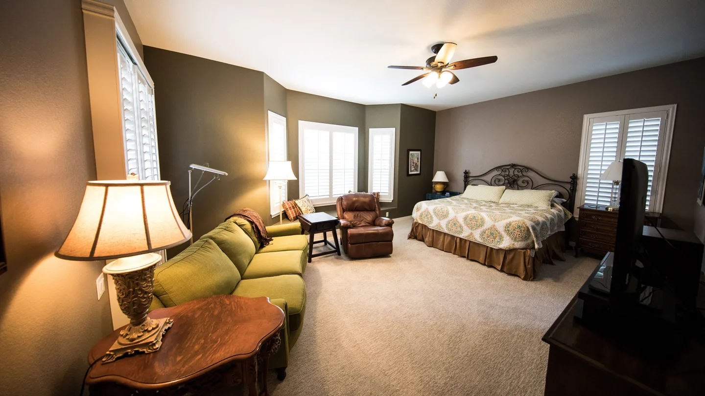 A master bedroom crowded with a wrought-iron bed, an olive sofa, a leather recliner, lamps and a television on a dresser